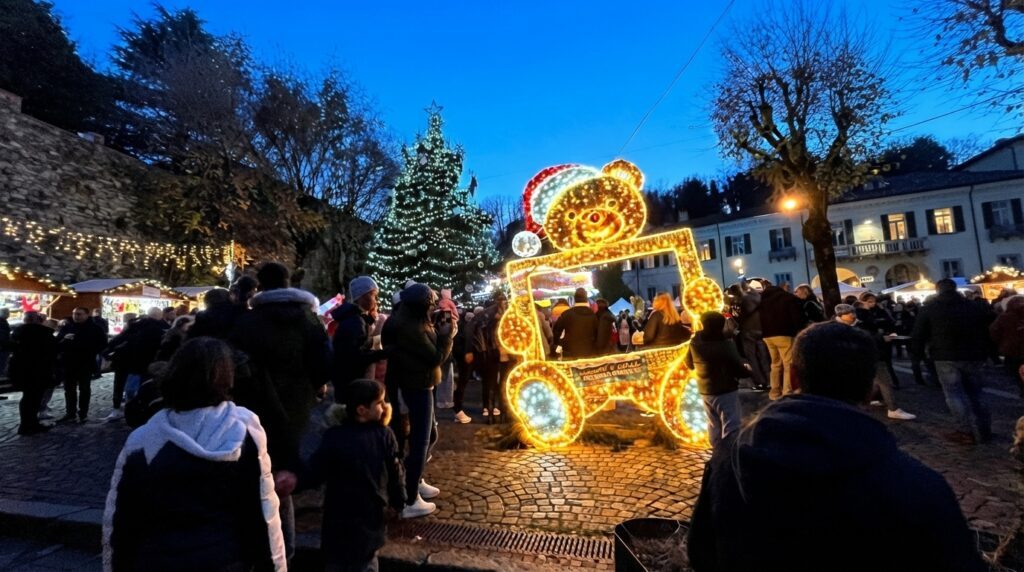 Una piazza affollata la sera con un grande albero di Natale illuminato e una luminosa installazione a forma di orsetto con cappello da Babbo Natale